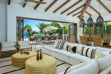 Bright beachfront villa open-plan living and dining area with vaulted wooden-beam ceiling, white sectional sofa and striped pillows, woven rattan ottomans and pendant lights, folding glass doors opening to a sunlit patio with umbrella, palm trees and ocean view.