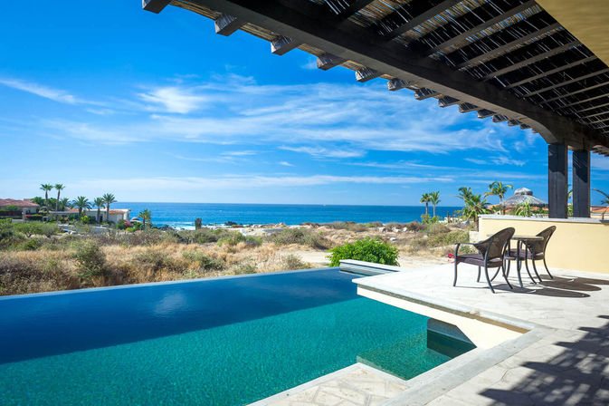 Sunny oceanfront infinity pool and shaded patio with two chairs overlooking sandy dunes, palm trees, and turquoise sea
