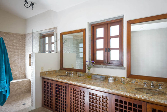 Bright Mediterranean-style bathroom with double-sink granite countertop, wooden lattice vanity, two wood-framed mirrors, small multi-pane window, mosaic-tiled walk-in shower with bench, and teal towel hanging