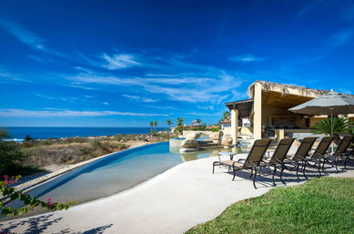 Oceanfront infinity pool with curved wading ledge, row of sun loungers and a thatched cabana overlooking the blue sea and sky.