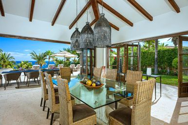 Sunlit open-air ocean-view dining area in a tropical villa with a glass-top table, woven rattan chairs, large wicker pendant lights, and sliding doors opening to a poolside patio, palm trees and blue sea.