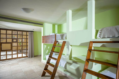Sunlit shared bedroom with pale green walls, built-in bunk niches holding white-bedded bunks accessed by wooden ladders, tiled floor and large wooden-framed glass doors with curtain