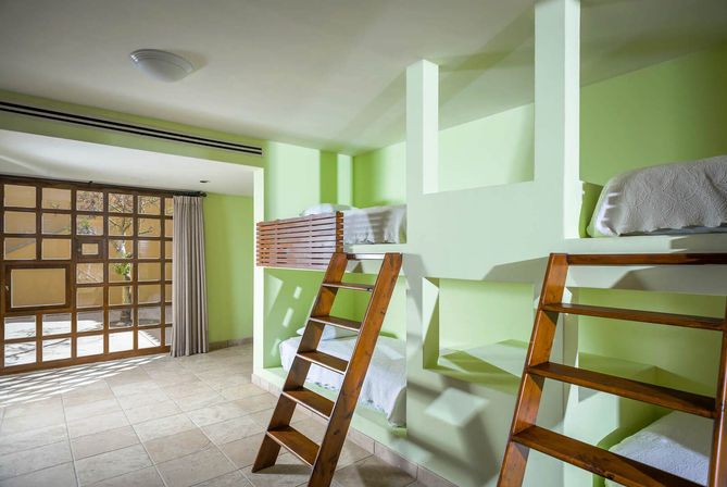 Sunlit shared bedroom with pale green walls, built-in bunk niches holding white-bedded bunks accessed by wooden ladders, tiled floor and large wooden-framed glass doors with curtain