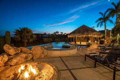 Evening tropical poolside at dusk with a lit palapa bar, infinity pool and circular spa, glowing fire pit, palm trees and lounge chairs under a deep blue sky.