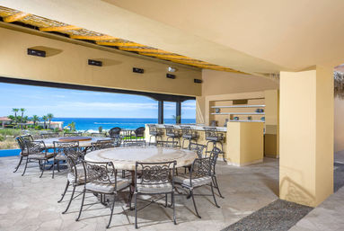 Oceanfront covered patio with round stone tables, decorative wrought-iron chairs and bar stools, tiled floor and open view of the blue sea and palm trees