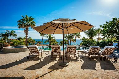 Sun-soaked oceanfront pool deck with cushioned chaise lounges under a large umbrella, palm trees and turquoise sea in the background