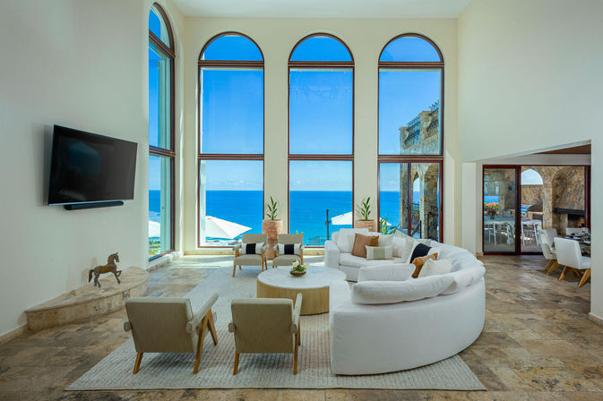 Sunlit luxury oceanfront living room with floor-to-ceiling arched windows framing turquoise sea, a white curved sectional, neutral armchairs, stone tile floors, wall-mounted TV and a seaside dining terrace visible through an archway.