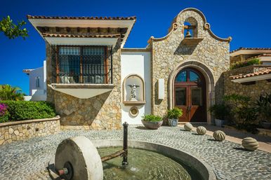 Sunlit Mediterranean-style stone villa entrance with arched wooden double doors, wrought-iron balcony, niche statue, cobblestone courtyard and round water fountain under a bright blue sky.