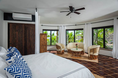 Sunny tropical coastal bedroom with white linens and blue patterned pillows, curved windows framing palm-tree views, cozy rattan seating, dark wood accents and terracotta tile floor.