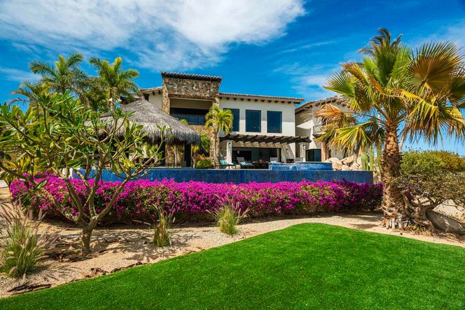 Sun-drenched tropical beachfront villa with stone and white stucco facade, infinity pool and thatched palapa, framed by palm trees and vibrant magenta bougainvillea under a bright blue sky.