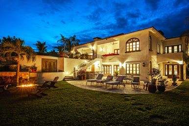 Luxury Mediterranean-style villa patio at dusk with warm exterior lighting, stone terrace and lounge chairs, fire pit, water feature, potted plants and palm trees