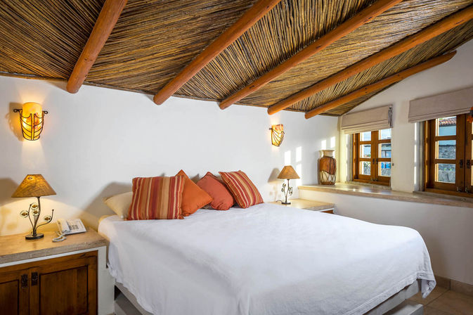 Cozy sunlit bedroom with white linens, orange striped pillows, bedside lamps and wooden windows under a woven reed ceiling with exposed beams — rustic Mediterranean-style cottage vibe.