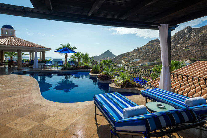 Oceanfront resort pool terrace with blue-striped lounge chairs, shaded cabana and umbrella, palm trees, tiled roofs and rugged coastal mountain views