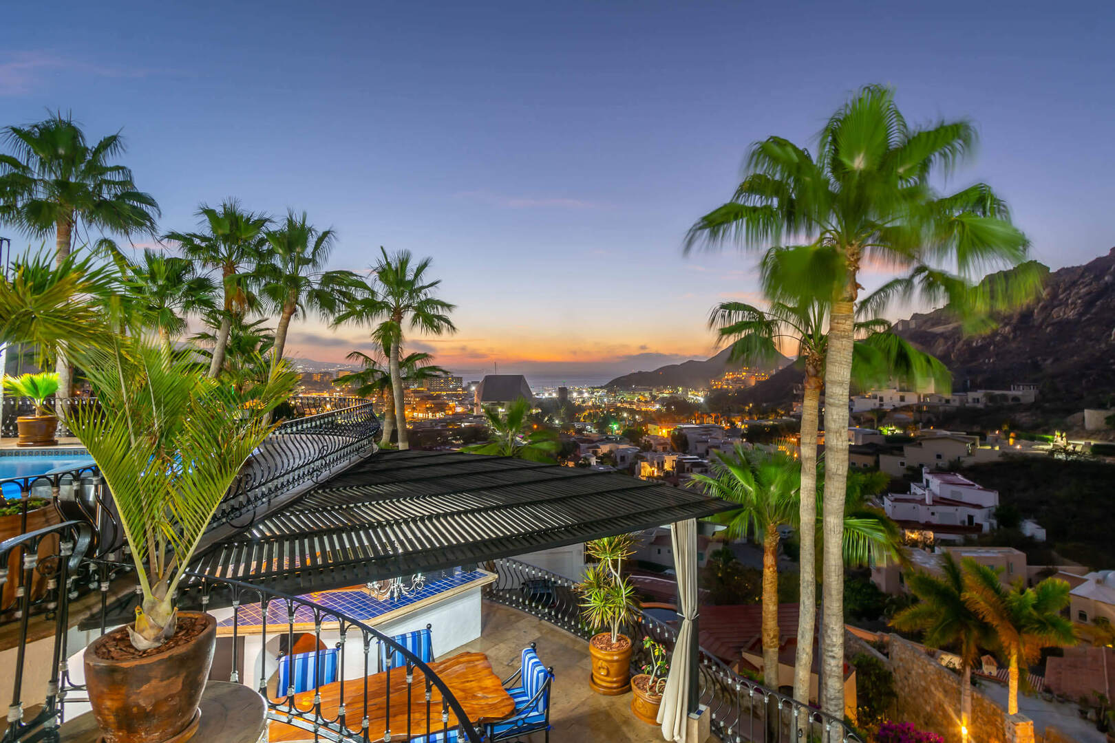 Rooftop terrace with pergola, potted palm trees and blue patio chairs overlooking a coastal town, marina and mountains at sunset with twinkling city lights.
