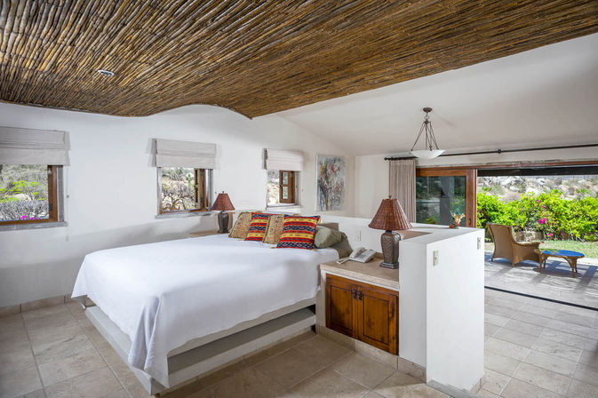 Sunny bedroom with rustic woven-wood ceiling, white platform bed topped with colorful patterned pillows, wicker bedside lamps, tiled floor and sliding glass doors opening to a lush garden patio.