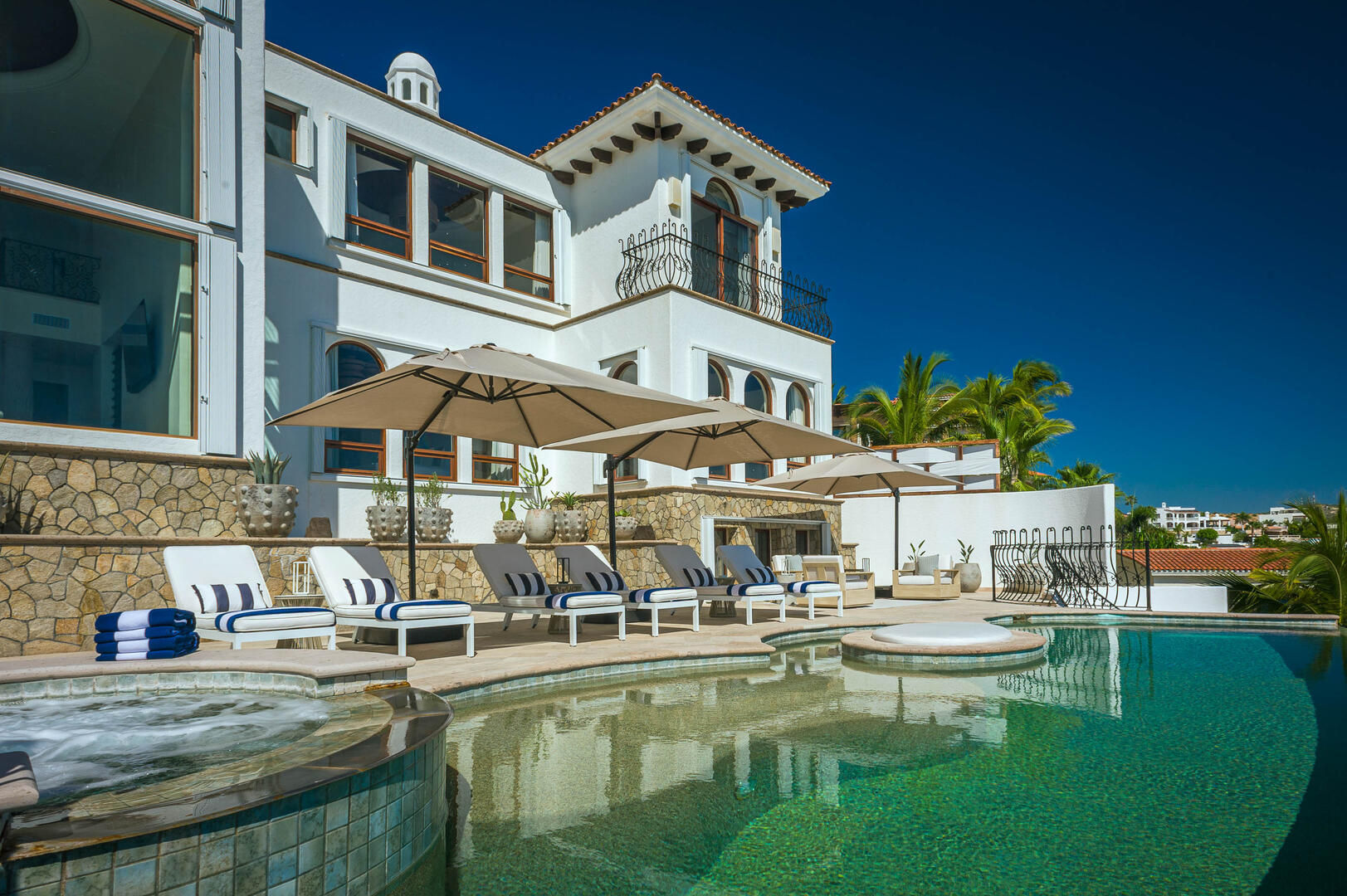 Sunlit Mediterranean-style villa with curved infinity pool, lined sun loungers with striped towels, large umbrellas, potted plants and palm trees under a deep blue sky.