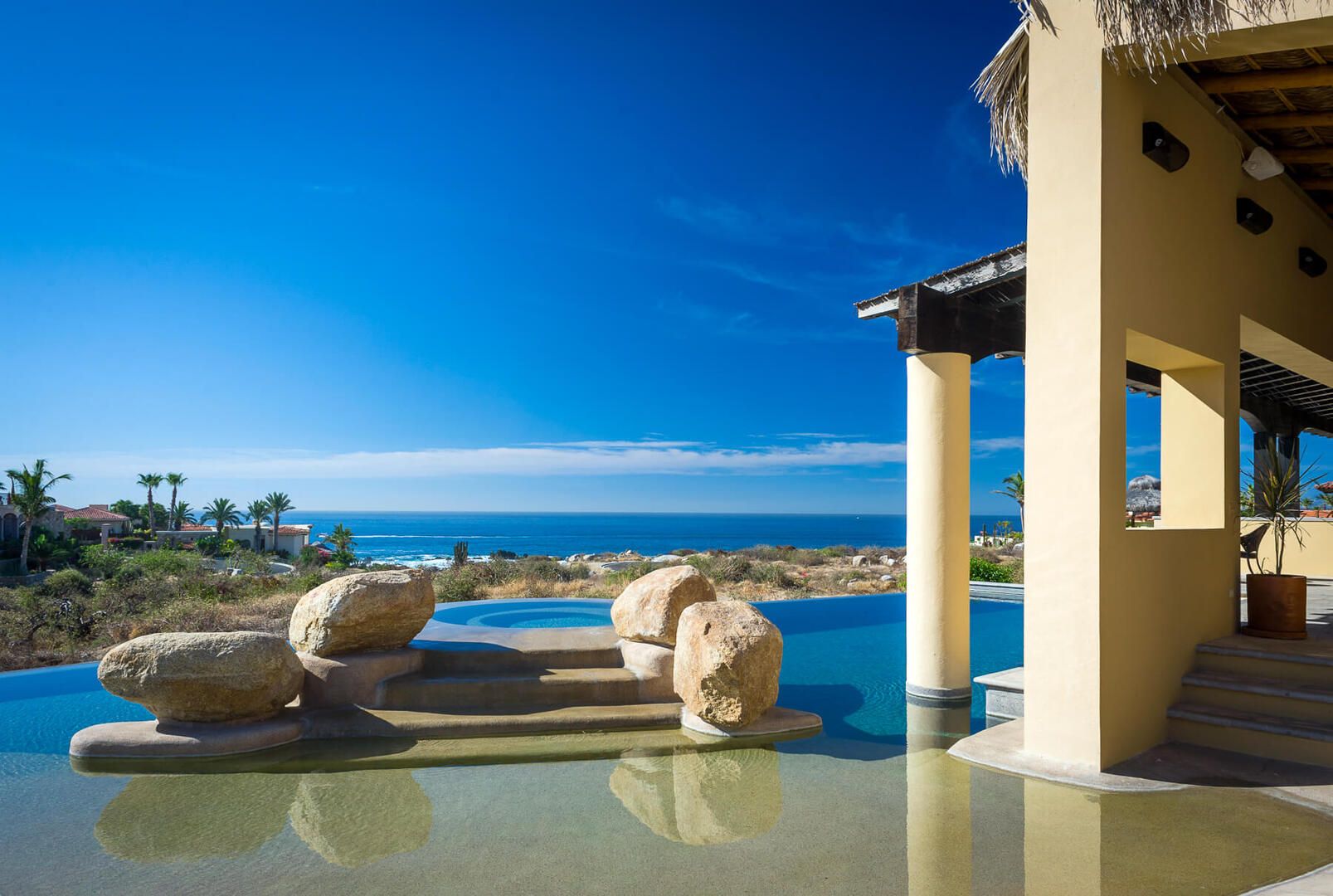 Sunlit oceanfront infinity pool at a coastal villa with large decorative boulders and steps into the water, shaded patio columns, palm trees and a clear blue sky over the coastline.