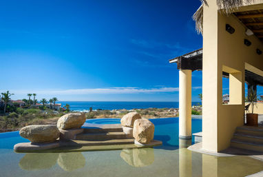 Sunlit oceanfront infinity pool at a coastal villa with large decorative boulders and steps into the water, shaded patio columns, palm trees and a clear blue sky over the coastline.
