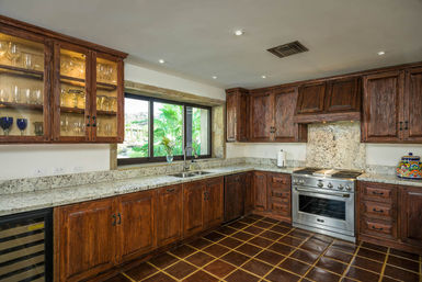 Cozy rustic kitchen with rich wood cabinets, speckled granite countertops, stainless steel range, glass-front cabinet with glassware, tile floor and leafy garden window view.
