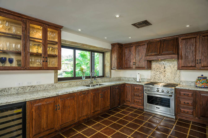 Cozy rustic kitchen with rich wood cabinets, speckled granite countertops, stainless steel range, glass-front cabinet with glassware, tile floor and leafy garden window view.