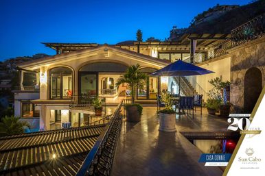 Evening view of a luxury hillside villa terrace in Los Cabos, Mexico — illuminated arches and balconies, potted palms, outdoor dining under a blue umbrella and a small reflecting pool.