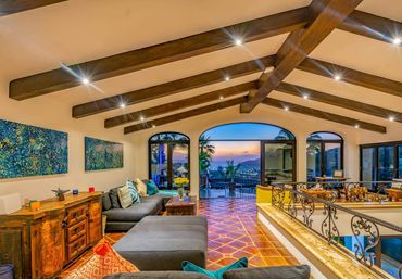 Mediterranean-style living room with exposed wooden beams, terracotta tile floor, gray sectional with colorful pillows, rustic wood sideboard and wrought-iron railing, opening through arched glass doors to a terrace and panoramic ocean sunset view.