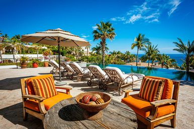 Sunny tropical ocean-view patio at a coastal resort with orange-cushioned wooden chairs around a rustic table, white chaise lounges under a large umbrella beside an infinity pool, palm trees and bright blue sky