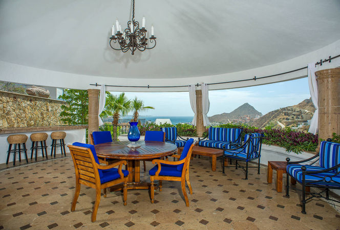 Coastal ocean-view outdoor dining terrace with round wooden table, blue-cushioned chairs, striped lounge seating, wrought-iron chandelier, bar stools and palm trees overlooking the sea and rocky hills.