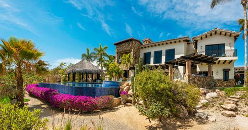 Luxury coastal villa with stone-and-white facade, infinity pool lined with bright pink bougainvillea, a thatched palapa swim-up bar, palm trees, cacti and a clear sunny blue sky.