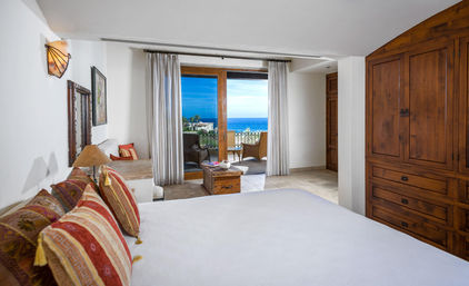 Cozy ocean-view bedroom with white bedding, patterned throw pillows, wooden armoire and sliding glass doors opening to a balcony with chairs and bright blue sea views.
