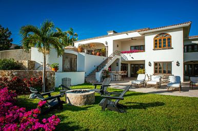 Sunny tropical Mediterranean-style villa with white stucco walls and terracotta roof, manicured lawn and palm tree, bright bougainvillea, stone fire pit surrounded by Adirondack chairs and spacious patio seating under a balcony.