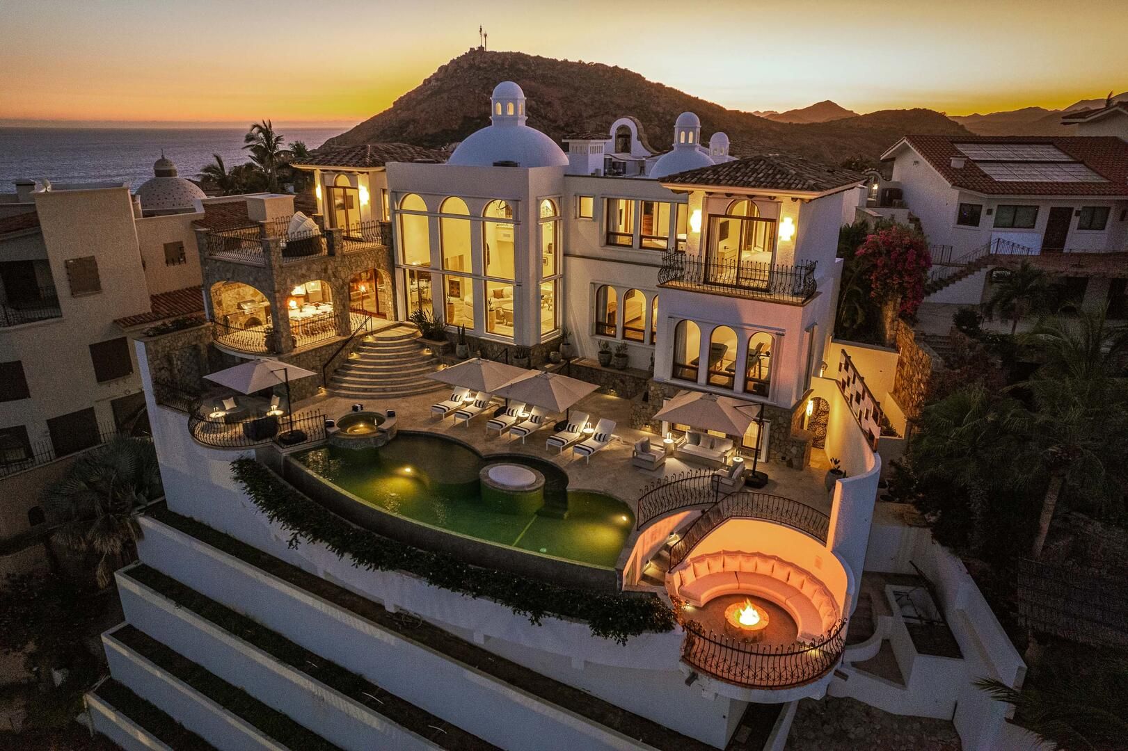 Sunset coastal hillside luxury villa with white Mediterranean domes, glowing windows, curved infinity pool, poolside loungers and umbrellas, and a sunken circular fire pit overlooking the ocean and distant mountains.
