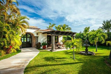 Sunny Mediterranean-style villa with arched doorway, shaded wooden pergola patio and dining set, lush tropical palms, manicured lawn and decorative stone fountain under a blue sky