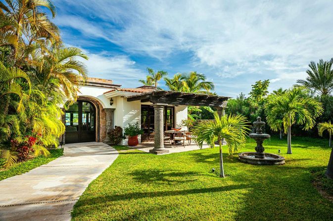 Sunny Mediterranean-style villa with arched doorway, shaded wooden pergola patio and dining set, lush tropical palms, manicured lawn and decorative stone fountain under a blue sky