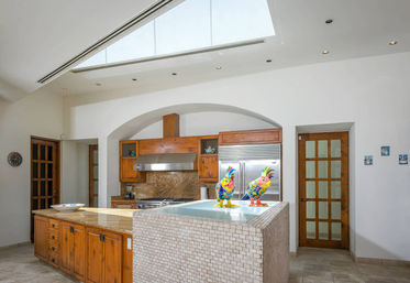 Bright modern kitchen with large skylight, arched alcove over warm wood cabinets, stainless steel range and refrigerator, granite countertops and mosaic-tiled island topped with two colorful ceramic rooster figurines.