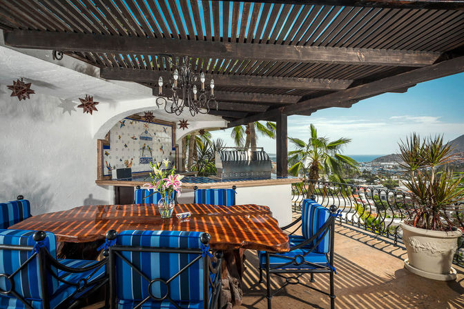 Sunlit ocean-view terrace with wooden pergola, rustic live-edge dining table, blue-striped chairs, chandelier and built-in grill, palm trees and coastal town visible beyond.