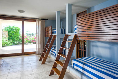 Bright coastal vacation bunk room with built-in wooden loft beds, blue-and-white striped mattresses and ladders, tiled floor, and sliding glass doors opening to a sunny garden with palm trees.