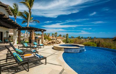 Sunny ocean-view tropical villa terrace with infinity pool and circular spa, wicker lounge chairs and palm trees under a bright blue sky