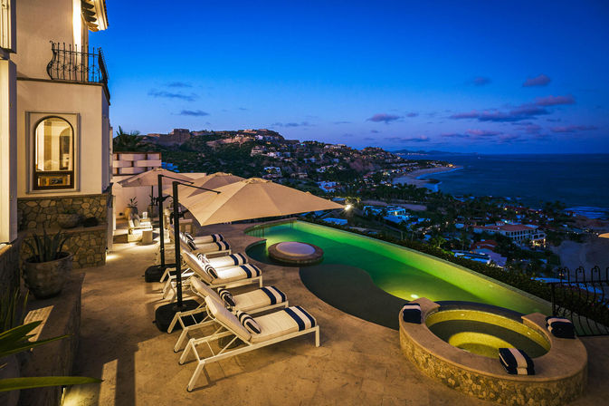 Twilight cliffside villa terrace with striped lounge chairs and umbrellas, glowing infinity pool and circular hot tub overlooking a panoramic coastal hillside and ocean shoreline.