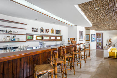 Sunlit tropical-style indoor bar with a polished wooden counter and bamboo bar stools, decorative wall shelves, small horizontal windows, textured thatch-style ceiling and tiled floor leading to a yellow sofa lounge.