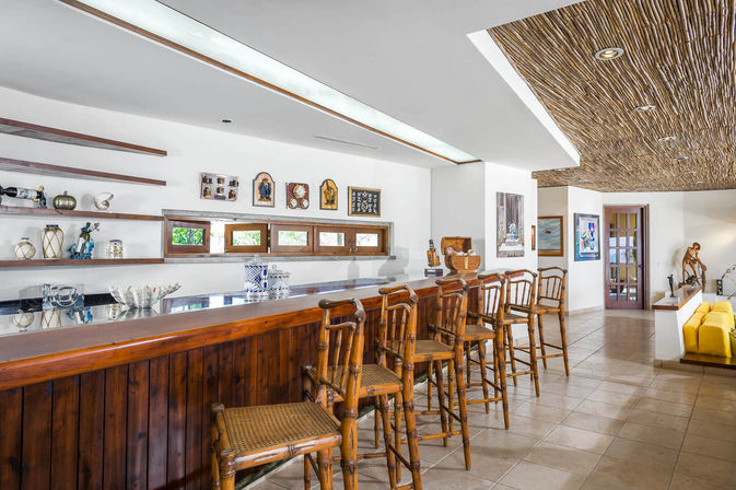 Sunlit tropical-style indoor bar with a polished wooden counter and bamboo bar stools, decorative wall shelves, small horizontal windows, textured thatch-style ceiling and tiled floor leading to a yellow sofa lounge.
