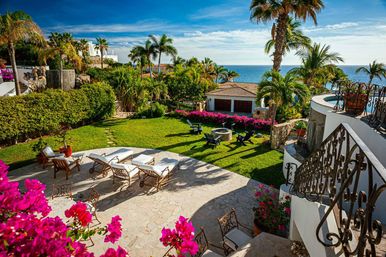 Sunny oceanfront villa terrace with palm trees, sun loungers on a stone patio, green lawn and fire-pit seating surrounded by vibrant bougainvillea and blue sea views.