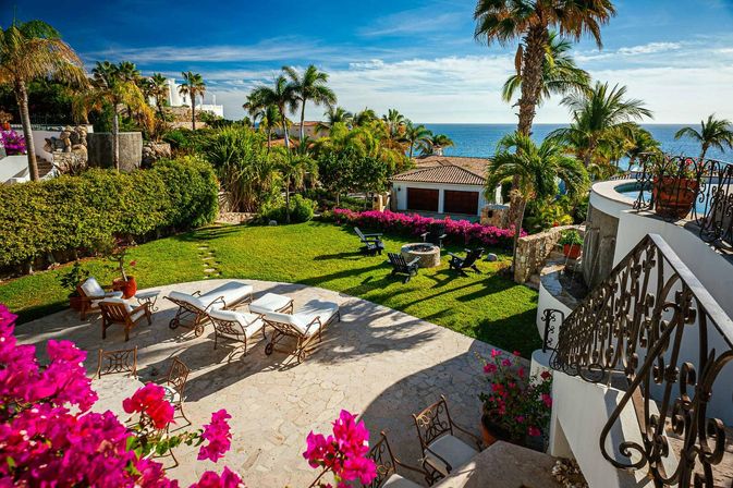 Sunny oceanfront villa terrace with palm trees, sun loungers on a stone patio, green lawn and fire-pit seating surrounded by vibrant bougainvillea and blue sea views.
