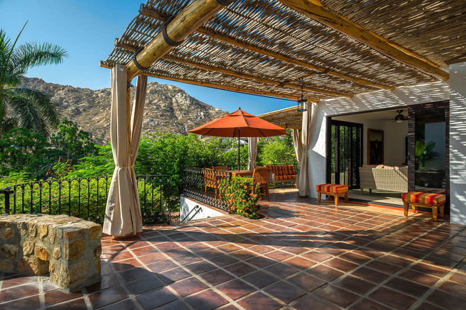 Sunny mountain-view terrace with rustic bamboo pergola, terracotta tile patio, orange umbrella and outdoor dining set surrounded by lush tropical greenery.