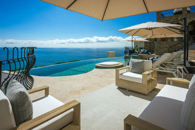 Sunlit oceanfront infinity pool terrace with cushioned wooden lounge chairs, umbrellas and a round plunge spa, overlooking the tranquil blue sea and horizon.