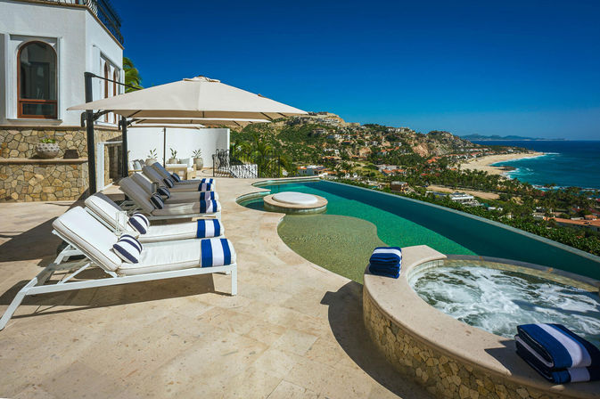Sunny oceanfront luxury villa terrace with an infinity pool and bubbling hot tub, lined with white sun loungers topped with blue-striped towels under a large umbrella, overlooking a sandy beach and rugged coastal hills.