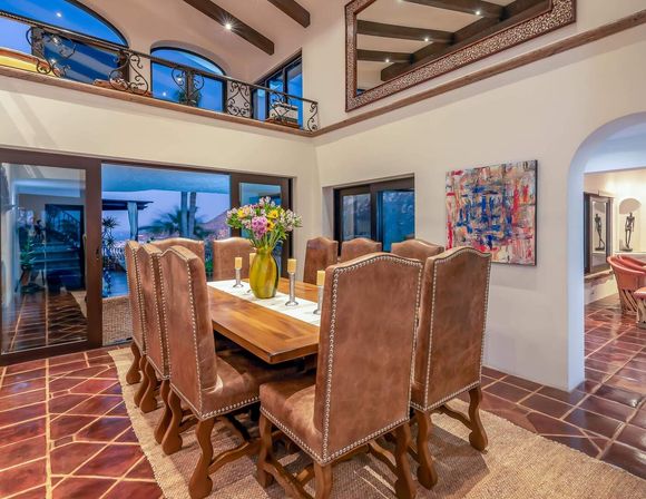 Spacious Mediterranean-style dining room with long wooden table and eight studded leather chairs, vase of fresh flowers centerpiece, terracotta tile floor, exposed wooden beams and wrought-iron balcony, sliding glass doors revealing a palm-lined coastal view.