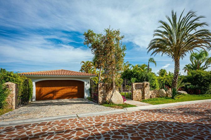 Sunlit Mediterranean-style house exterior with terracotta roof and wooden garage door, gated stone wall and cobblestone driveway surrounded by palm trees and lush tropical landscaping under a blue sky