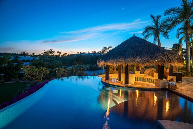 Tropical infinity pool at dusk with an illuminated thatched palapa swim-up bar, palm trees silhouetted against a crescent moon and colorful sky, and warm reflections on calm water.
