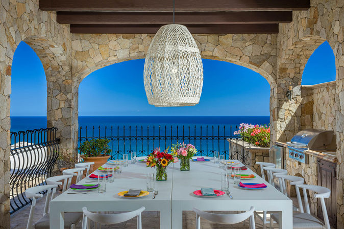 Sunny oceanfront alfresco dining terrace with Mediterranean-style stone archways, long white table set with colorful plates and flower vases under a woven pendant lamp, wrought-iron railing and built-in grill overlooking the deep blue sea